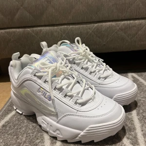 Vita chunky sneakers från Fila - Säljer ett par vita Fila sneakers med chunky sula och holografiska detaljer på sidan och plösen. Skorna har snörning, rund tå och är tillverkade i syntetmaterial med meshfoder för extra komfort. Perfekt för dig som gillar en sportig och trendig look.