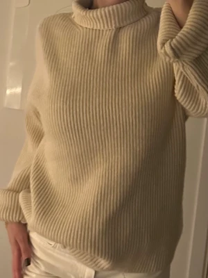 Beige ribbad polotröja i ull - Säljer en beige polotröja med ribbad struktur och hög krage. Tröjan är stickad i mjuk ull och har en loose passform med långa ärmar. 