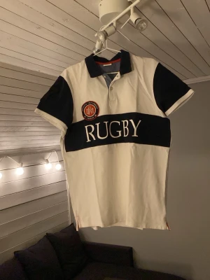 Vit och marinblå t-shirt - Snygg t-shirt i vit och marinblå med klassisk krage och knappslå. Stor text 'RUGBY' över bröstet och ett broderat märke på vänster sida. Perfekt för dig som gillar sportig stil och vill sticka ut.