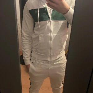 Vit och grön dress från Lacoste - Säljer en fräsch vit dress från Lacoste med grönt block över bröstet och sidorna. Tröjan har huva med snören, dragkedja framtill och två fickor. Klassisk Lacoste-logga på bröstet och detaljer på dragkedjan. Perfekt för en sportig och chill look.