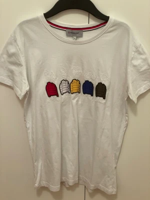 Vit Moncler t-shirt med maya broderi - Vit t-shirt från Moncler med coolt broderi på bröstet som visar fem färgglada dunjackor och tecknade ansikten. Klassisk Moncler-logga på ärmen. T-shirten har rund hals och korta ärmar, tillverkad i mjuk bomull. Perfekt för dig som gillar street och designer.