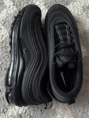 Nike Air Max 97 Triple Black 40.5 - Mycket  gott skick  AirMax 97 Triple Black med vågiga paneler och synlig Air-sula. Helt svarta sneakers med mesh och syntetmaterial, snörning och reflekterande detaljer. Ikonisk design som är både sportig och stilren, perfekt för dig som gillar streetwear.