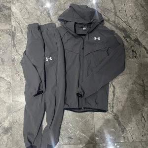 Grå träningsset från Under Armour - Säljer ett grått träningsset från Under Armour i storlek M. Setet består av en hoodie med dragkedja och matchande byxor, båda med logga på bröst och ben. Materialet är 87% nylon och 13% spandex, vilket gör det lätt och flexibelt. Perfekt för löpning och sport.