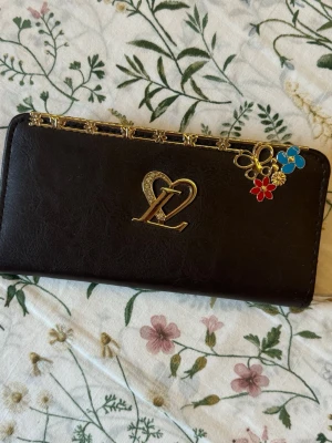Plånbok med gulddetaljer och blommor - • Brun plånbok i skinn med guldfärgad dekor upptill och ett hjärtformat emblem på framsidan.  • Plånboken har dragkedja med hjärtformad detalj och små blommor i guld samt färgglada blomcharms i hörnet.  • Perfekt för kort och kontanter. • Är trasig på vissa ställen som det ses i bilderna. 