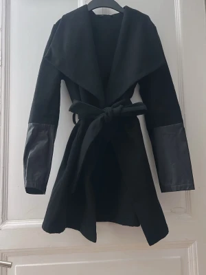 Svart omlottkappa med skinnärmar - Svart kappa med bred krage och omlottmodell, knyts i midjan med bälte. Ärmarna har snygga detaljer i svart skinn som ger en cool kontrast till resten av kappan som är i ull. Perfekt för dig som gillar stilrena och moderna plagg.