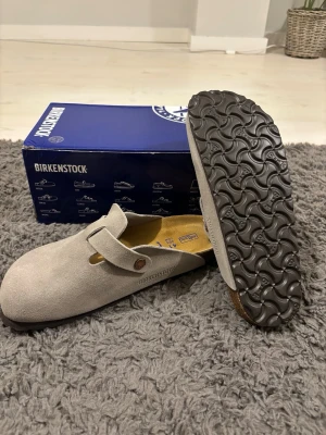 Birkenstock Boston grå mocka  - Birkenstock Boston i grå mocka med rund tå och platt sula. Skorna har justerbar rem med metallspänne och mjuk fotbädd för extra komfort. Perfekt för dig som gillar klassisk stil och vill ha något riktigt bekvämt. Levereras med originalkartong och tags. Har aldrig används utomhus bara runt hemmet. Passar storlek 40.