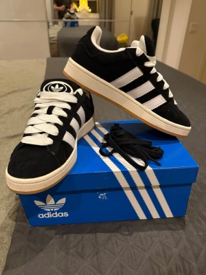 Adidas Campus sneakers svart/vit - Säljer ett par Adidas Campus sneakers i svart mocka med vita detaljer och klassiska tre ränder på sidan. Skorna har vit platt sula och rund tå, samt snörning med vita skosnören. Gummisulan är ljusbrun och ger bra grepp. Box och extra svarta skosnören medföljer.