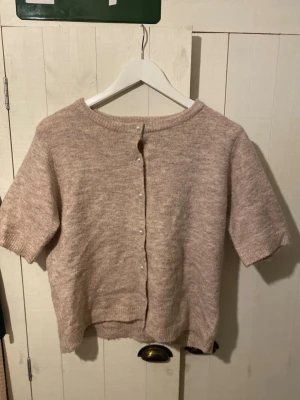 Beige stickad kofta från 157 - Kortärmad beige stickad kofta från 157 i storlek M. Koftan har rund hals och knappar framtill, perfekt för lager-på-lager. Mjuk och skön känsla med ribbade kanter vid hals och ärmslut. Enkel och stilren design som funkar året runt.