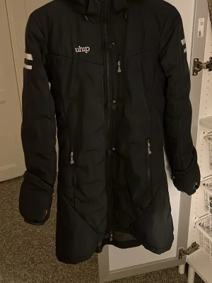 Ridkappa från Uhip - Marinblå parkajacka från Uhip med orange foder, huva och flera praktiska dragkedjor. Jackan har reflexdetaljer på ärmar och rygg, samt fickor med dragkedja. Perfekt för kalla dagar och har en stilren look med lång passform.