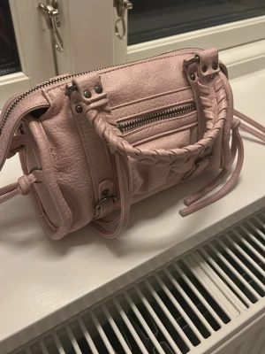 Ljusrosa handväska med flätade handtag - Supersöt ljusrosa handväska i skinnimitation med flätade handtag och coola metalldetaljer. Väskan har dragkedja upptill, en liten ficka med dragkedja framtill och långa dekorativa remmar på sidorna. Perfekt storlek för det viktigaste och ger en trendig touch till din outfit. En remsa har släppt (se sista bild) men ingenting man tänker på!🥰