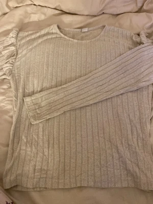 Beige ribbad tröja med volang från Lindex - Säljer en beige långärmad tröja från Lindex i storlek 170. Tröjan har ribbat mönster och snygga volangdetaljer på axlarna som ger en cool touch. Perfekt för dig som gillar stilrena plagg med lite extra detaljer.