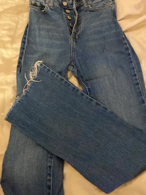 Blå wide jeans från Trendyol, strl 34 - Säljer ett par blå jeans från Trendyol i storlek 34. Modellen är utsvängd/bootcut med hög midja och råa, fransiga benslut. Klassisk knappgylf framtill. .