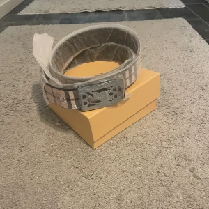 Rutig Burberry bälte med silverdetalj - Säljer ett snyggt rutig bälte från Burberry med klassiskt mönster i beige, grått och brunt. Bältet har en stilren silverfärgad spänne med Burberrys logotyp. Perfekt för att lyfta din outfit med en lyxig touch.