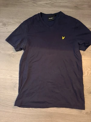 Marinblå t-shirt från Lyle & Scott - Snygg marinblå t-shirt från Lyle & Scott med gul broderad logga på bröstet. T-shirten har v-ringad hals och korta ärmar, tillverkad i mjuk bomull som känns skön mot huden. Perfekt för dig som gillar stilrena och klassiska plagg.
