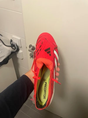 Adidas Predator elite  - Säljer ett par Adidas Predator fotbollsskor i röd färg med vita och svarta detaljer. Skorna har snörning, spetsig tå och platt sula med dobbar för konstgräsplan. Ovandelen är i syntetmaterial och har coola grafiska linjer på sidorna. Perfekt för dig som vill sticka ut på planen. Säljes då dom inte passar som tänkt