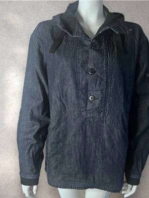 Stone Island jeansjacka med huva - Snygg mörkblå jeansjacka från Stone Island med huva och svarta snören. Jackan har tre knappar framtill och klassisk Stone Island patch på vänster ärm. Långärmad och loose fit, perfekt för dig som gillar streetwear. Plagget har inga defekter och inga revor. Till och med det inre fodret är i perfekt skick. Det finns bara ett lätt slitage på ärmen, som knappt är märkbart och inte påverkar användningen på något sätt.#stoneisland #jeansjacka