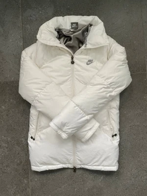 Vit pufferjacka från Nike  - Snygg vit pufferjacka från Nike Sportswear med broderad logga på bröstet och coolt metallmärke på dragkedjan. Jackan har hög krage, två sidofickor med dragkedja och ett silvrigt innerfoder. Perfekt för kalla dagar och riktigt stilren look. Storleken är S men jag skulle säga att den sitter lutar sig lite mer mot XS hållet. Nypris: 1200kr                                  Pris går att diskutera☺️