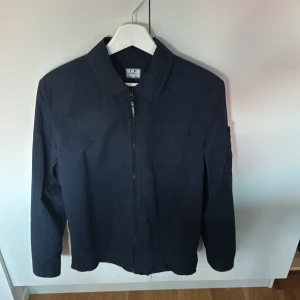 Marinblå overshirt från C.P. Company - Säljer en marinblå overshirt från C.P. Company med dragkedja framtill och klassisk krage. Jackan har en ficka på bröstet och den ikoniska linsen på ärmen. Tillverkad i lätt polyester, perfekt för lager på lager och streetwear-vibe.