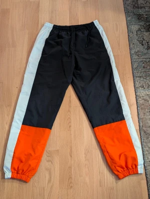 Färgglada träningsbyxor från Lacoste - Säljer ett par snygga träningsbyxor från Lacoste i storlek M. Byxorna har cool färgblock-design med svart, ljusgrå och orange, elastisk midja med snörning och meshfoder. Perfekt för dig som gillar sportig och trendig stil.