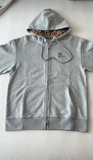 Grå hoodie från Burberry med dragkedja - Snygg grå hoodie från Burberry med klassiskt rutigt foder i huvan och broderad logga på bröstet. Tröjan har snörning i huvan, två fickor fram och hel dragkedja. Perfekt för dig som gillar stilrena detaljer och exklusiva märken.