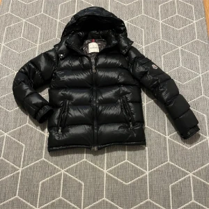 Svart Moncler pufferjacka med huva - Svart pufferjacka från Moncler med glansig yta, huva och två dragkedjeförsedda fickor. Jackan har Moncler-logga på ärmen och coolt serietryck på insidan. Perfekt för dig som vill ha en varm och snygg jacka. Kan skicka code moncler länken vid önskan.