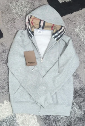 Grå hoodie från Burberry med dragkedja - Grå hoodie från Burberry i mjuk bomull med klassisk rutig insida på huvan. Tröjan har snörning vid huvan, dragkedja framtill och subtilt broderad Burberry-logga på bröstet. Perfekt för dig som gillar stilrena och lyxiga detaljer.