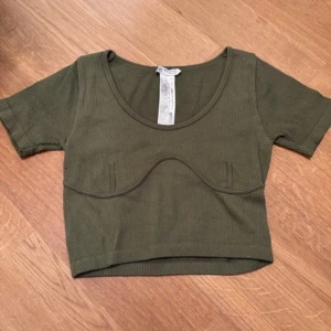 Olivgrön ribbad croppad topp - Snygg olivgrön croppad topp från H&M med ribbad struktur och markerad söm under bysten. Rundad halsringning och korta ärmar ger en cool och modern vibe. Perfekt att styla med höga jeans eller kjol för en trendig look.