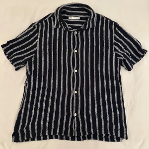 Randig kortärmad skjorta från Zara - Snygg kortärmad skjorta från Zara i storlek L med vertikala vita och mörkblå ränder. Skjortan har klassisk krage, vita knappar framtill och är tillverkad i mjuk bomull och polyesterblandning. Perfekt för dig som gillar stilrena och moderna mönster.
