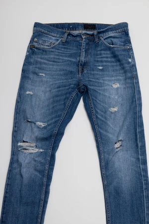 Blå slitna jeans från Tiger of Sweden  - Snygga blå jeans från Tiger of Sweden med slitningar och hål på framsidan för en cool, avslappnad vibe. Klassisk femficksmodell med raka ben och normal passform. Jeansen har en medelhög midja och är gjorda i mjukt denimtyg med lätt tvättad look.