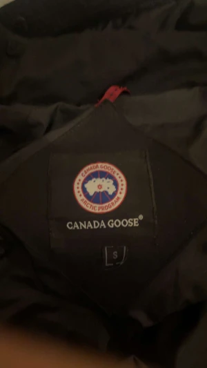 Svart dunjacka Canada Goose S - Svart dunjacka från Canada Goose i storlek S. Jackan har huva, dragkedja och knappar framtill, samt två fickor med dragkedja. Klassisk patch på ärmen och snygg, stilren look som håller dig varm hela vintern.