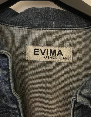 Mörkblå jeansbolero från Evima - Snygg mörkblå jeansbolero från Evima med korta ärmar, dubbla fickor framtill och bred krage. Jackan har dekorativa sömmar över bröstet och är croppad i modellen.