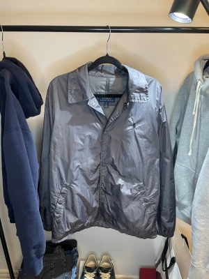 Grå vindjacka från Polo Ralph Lauren - Snygg grå vindjacka från Polo Ralph Lauren med klassisk krage och knäppning framtill. Jackan har diskret broderad logga på bröstet och två sidofickor. Tillverkad i glansig polyester som ger en clean och modern look.