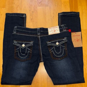 True Religion Ricky Super QT jeans, mörkblå - Va inte blyga med att pruta eller fråga om mer information. :). Mörkblå jeans från True Religion, modell Ricky Super QT. Snygga kontrastsömmar i vitt och brunt, klassiska fickor med lock och silverknappar. Märkespatch baktill och detaljerad brodering på bakfickorna. Slim passform och tillverkade i jeansmaterial.
