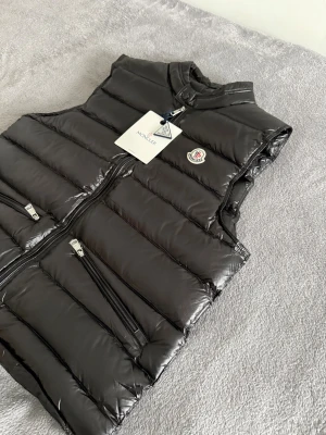 Moncler väst - Snygg svart dunväst från Moncler med glansig finish och quiltad design. Västen har ståkrage, två dragkedjeförsedda fickor och Moncler-logga på bröstet. Perfekt för lager-på-lager och streetstyle. Dragkedja framtill och diskret knappdetalj vid halsen.