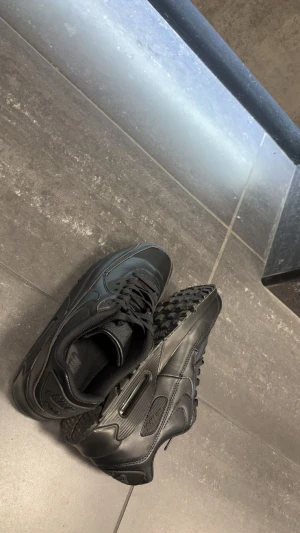 Svarta Nike Air Max sneakers - Säljer ett par helsvarta Nike Air Max 90 sneakers med klassisk design. Skorna har en chunky sula, snörning och är tillverkade i skinn och syntet. Perfekta för dig som gillar en clean och sportig look.