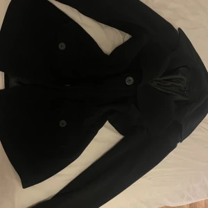 Svart kappa från Vila i ullmix - Stilren svart kappa från Vila med klassisk krage och stora knappar framtill. Jackan har två fickor med lock och är helfodrad. Perfekt för dig som vill ha en tidlös och snygg look under kyliga dagar.