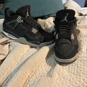 Svarta Nike Jordan 4 sneakers - Säljer ett par svarta Nike Jordan 4 sneakers med grå och vita detaljer. Skorna har klassisk snörning, meshpaneler och en robust sula. Ikonisk Jumpman-logga på plösen och cool design som sticker ut. Perfekta för dig som gillar streetwear och vill ha något unikt.
