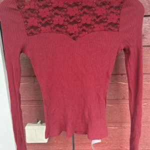 Röd ribbad långärmad topp från FASISTER - Snygg röd långärmad topp från FASISTER i storlek XS. Toppen har ribbad struktur och en vacker spetsdetalj med blommönster över bröstet. Figurnära passform och rund halsringning. Perfekt för dig som gillar detaljer och vill sticka ut.
