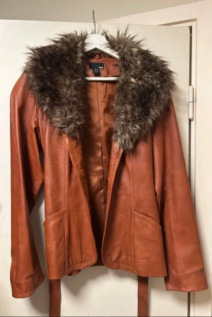 Brun skinnjacka med fuskpäls från H&M - Jacka i äkta skinn från H&M med fuskpälskrage. Kan knäppas med tre knappar samt ett band rund midjan, har även två fickor fram. Kragen går att ta av med knappar.