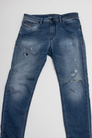 Diesel blå slitna jeans - Blå jeans från Diesel med snygga slitningar och lagade hål på framsidan. Modellen har tapered fit, klassiska fem fickor och knappgylf. Jeansen har en cool tvättad look och detaljer som förstärkta sömmar och Diesel-logga på bakfickan.