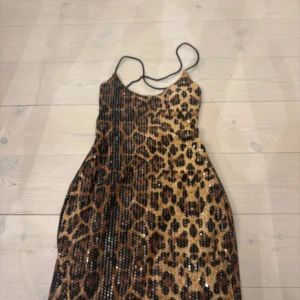 Leopardmönstrad paljettklänning - Snygg kort klänning med leopardmönster och glittriga paljetter. Klänningen har tunna axelband, snörning i ryggen och figurnära passform. Perfekt för dig som vill sticka ut med ett djurmönstrat statement-plagg.