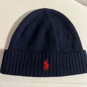 Marinblå merinoullsmössa Polo Ralph Lauren - Snygg marinblå mössa från Polo Ralph Lauren i mjuk och tvättbar merinoull. Mössans storlek är i One size. Klassisk ribbad kant och röd broderad logga framtill. Perfekt för dig som vill ha en stilren och varm mössa med premiumkänsla. Vid frågor är det alltid att höra av sig! 