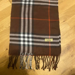 Burberry halsduk - Exklusiv halsduk från Burberry i 100% kashmir, tillverkad i England. Snyggt rutmönster i brunt, vitt, svart och orange med klassiska fransar i ändarna. Perfekt accessoar för att lyfta din stil med lyxig känsla och tidlös design.