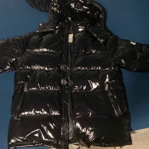 Svart pufferjacka från Moncler - Säljer en svart, glansig pufferjacka från Moncler med huva och dragkedja framtill. Jackan har två fickor med dragkedja och Moncler-logga på ärmen. Materialet är syntet och dun, perfekt för kalla dagar. Jackan har en clean och modern look som sticker ut.