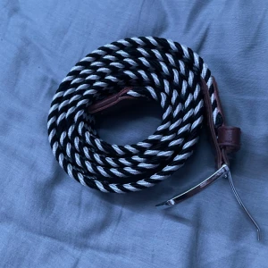 Randigt svartvitt flätat bälte - Säljer ett snyggt flätat bälte med svartvita ränder och bruna skinn-detaljer vid spännet. Bältet har ett coolt tvinnat mönster och ett silvrigt metallspänne som ger en unik look. Perfekt för att lyfta din outfit med lite extra edge.