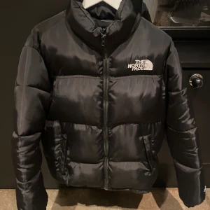 Svart pufferjacka från The North Face - Säljer en svart pufferjacka från The North Face i storlek M. Jackan har broderad logga på bröstet och ryggen, hög krage och dragkedja framtill. Materialet är lätt glansigt och känns riktigt mjukt, perfekt för kalla dagar. Jackan har två sidofickor och en clean, stilren look. Minimal skavank på höger ficka då en kort söm har gått upp, enkelt att fixa om man så skulle vilja.