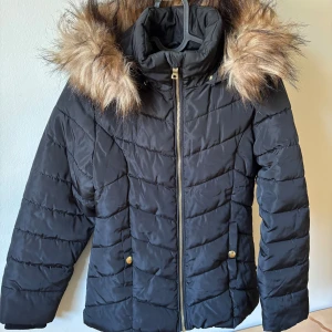 Svart jacka med fuskpäls - Svart pufferjacka med huva och snygg fuskpäls runt kragen. Storlek 140.