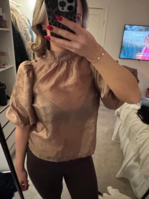 Champagnefärgad blus med puffärm - Säljer en champagnefärgad blus med högt halslinje och puffiga korta ärmar. Blusen har en lätt glansig yta och är tillverkad i ett tunt, elegant material som ger en lyxig känsla. Perfekt för dig som vill sticka ut med en trendig och chic look.