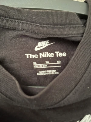 Svart Nike t-shirt i bomull XL (barn) - Svart t-shirt från Nike i storlek XL, med klassisk vit Nike-logga på bröstet. Modellen har rund hals och korta ärmar, tillverkad i mjuk bomull som är skön mot huden. Perfekt för dig som gillar enkel och sportig stil.