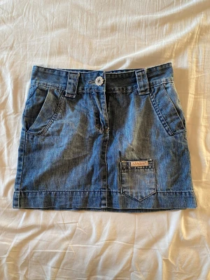 Blå jeanskjol från Vingino - Snygg blå jeanskjol från Vingino med coola fickor fram och bak, bälteshällor och en blomformad knapp. Kjolen har broderad etikett med fjärilar och Vingino-logga. Perfekt för dig som gillar denim och detaljer. Vintage, storlek 30 i midja men liten 